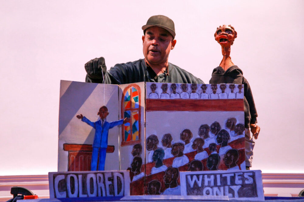 Schroeder Cherry, dressed in all black and wearing a blaseball hat, stands behind a large folded cardboard scene with one of his handmade puppets. In the scene, a preacher in a blue suit, arms outstretched above the word, "colored," faces rows of seated individuals in a full church above the words, "whites only."