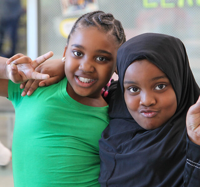 Two ASALA students — one in a green shirt and one in a black hijab — smile and make peace signs at the camera.