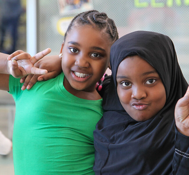 Two ASALA students — one in a green shirt and one in a black hijab — smile and make peace signs at the camera.