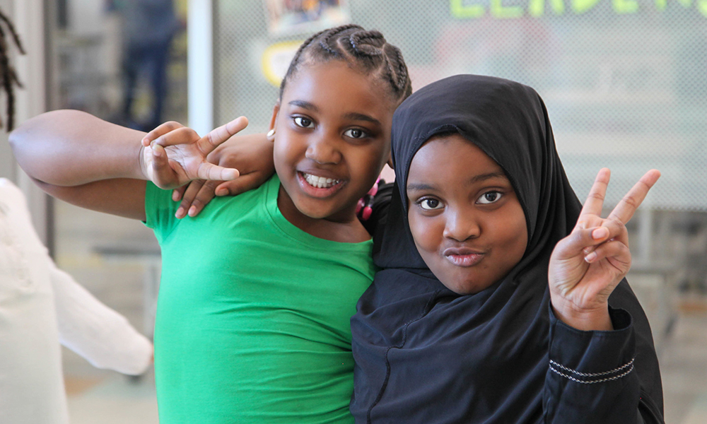 Two ASALA students — one in a green shirt and one in a black hijab — smile and make peace signs at the camera.