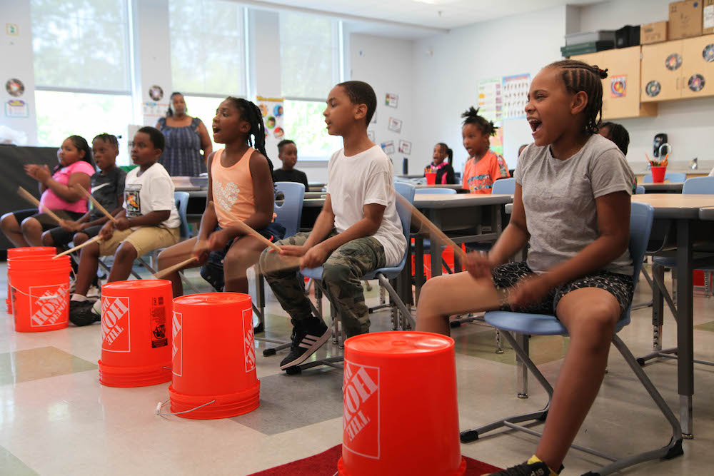 Students are dressed for warm weather in t-shirts and shorts. They are in a classroom, lined up on chairs, and drumming on overturned orange buckets.