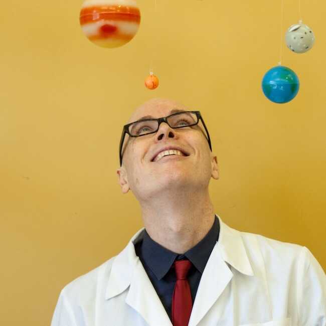 Dino O'Dell wears a white lab coat and gazes up towards model planets floating in space around him. He and the planets are against a yellow ochre background.