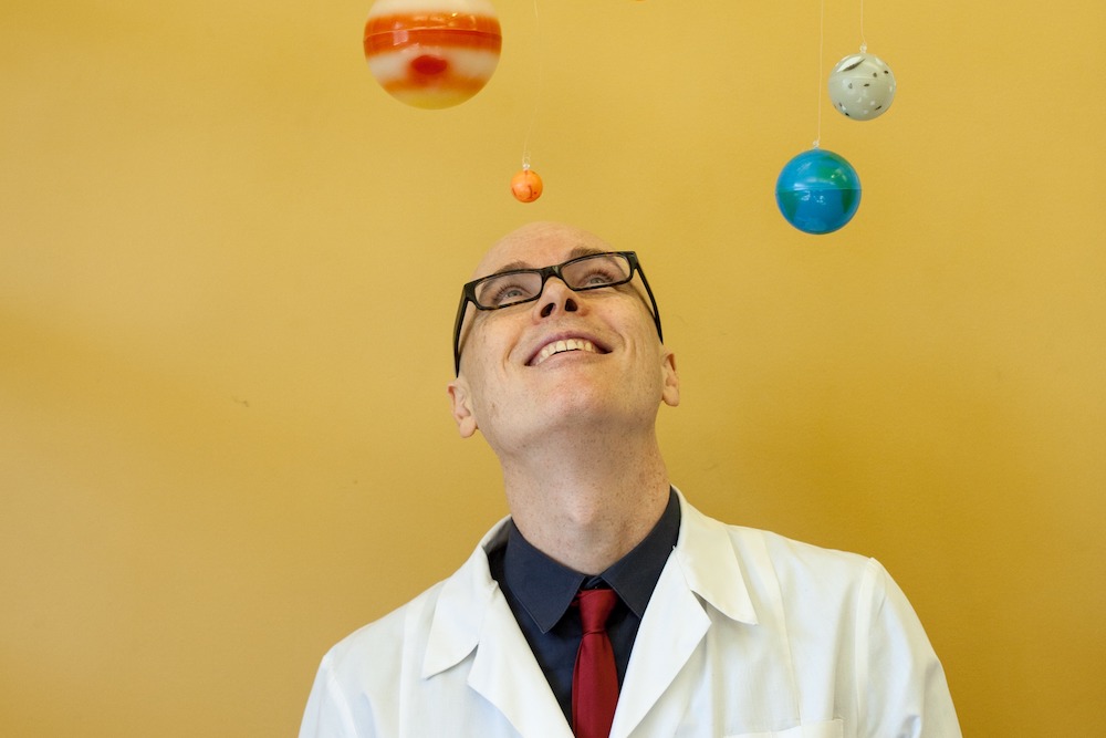 Dino O'Dell wears a white lab coat and gazes up towards model planets floating in space around him. He and the planets are against a yellow ochre background.