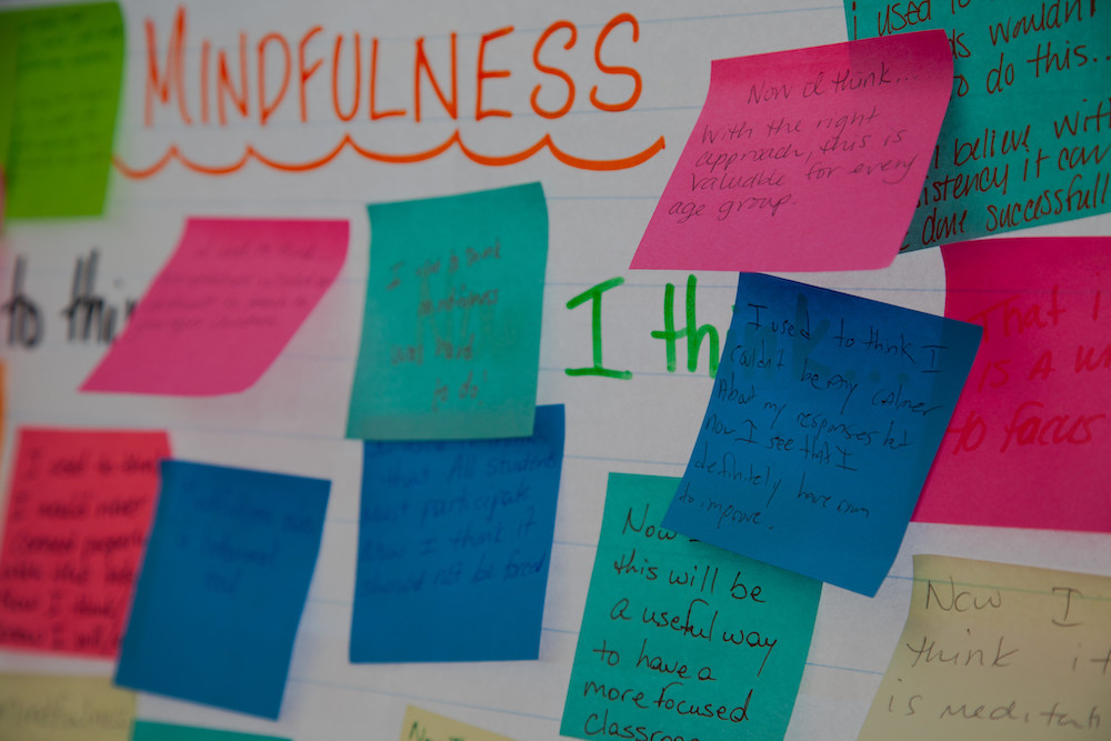 Pink, green, and blue post-it notes cover a white board. The word 'mindfulness' is written at the top of the board in red letters. The post-it notes have the handwriting of many people and convey their thoughts about mindfulness.