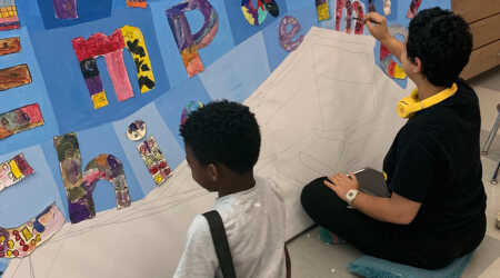Students work on a painted mural with letters on a blue background.