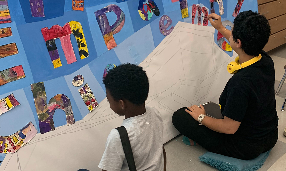 Students work on a painted mural with letters on a blue background.