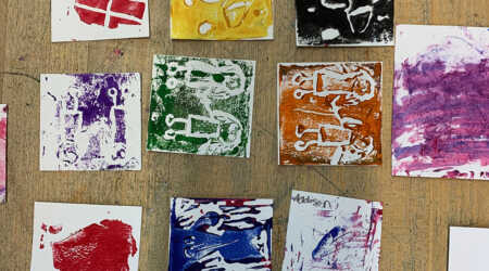 Square student monoprints on paper lie on a wooden table.