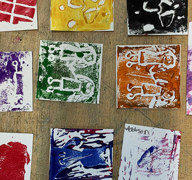 Square student monoprints on paper lie on a wooden table.