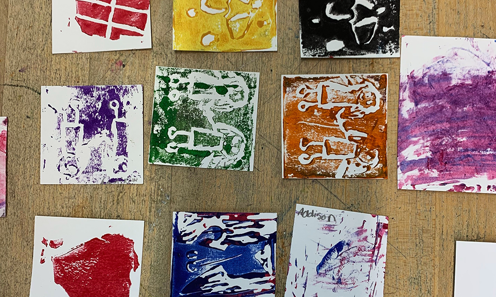Square student monoprints on paper lie on a wooden table.
