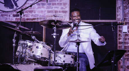 Vinton Jones smiles and speaks into a microphone while sitting at a drum kit.