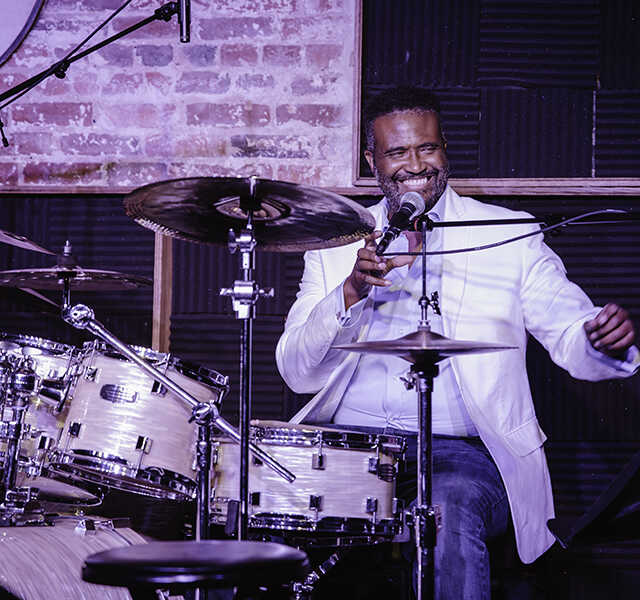 Vinton Jones smiles and speaks into a microphone while sitting at a drum kit.