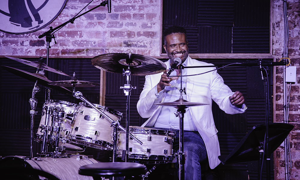 Vinton Jones smiles and speaks into a microphone while sitting at a drum kit.