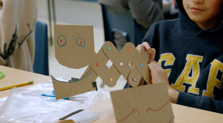A student holds a creature created with cardboard pieces and colored brads.