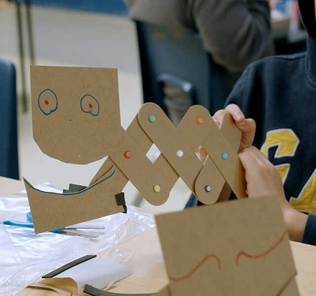 A student holds a creature created with cardboard pieces and colored brads.