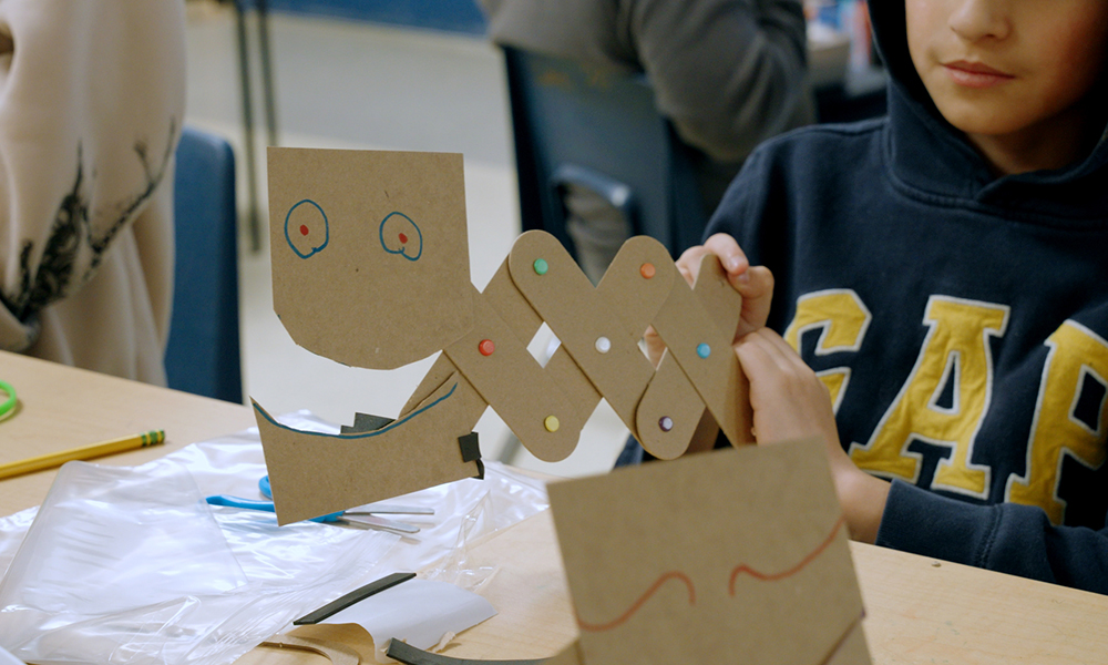 A student holds a creature created with cardboard pieces and colored brads.