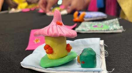 a small, yellow polymer clay fairy house with a pink, pointed roof sits next to a tiny green figure atop a hand-sewn cloth pocket. A students hands can be seen working in the background.