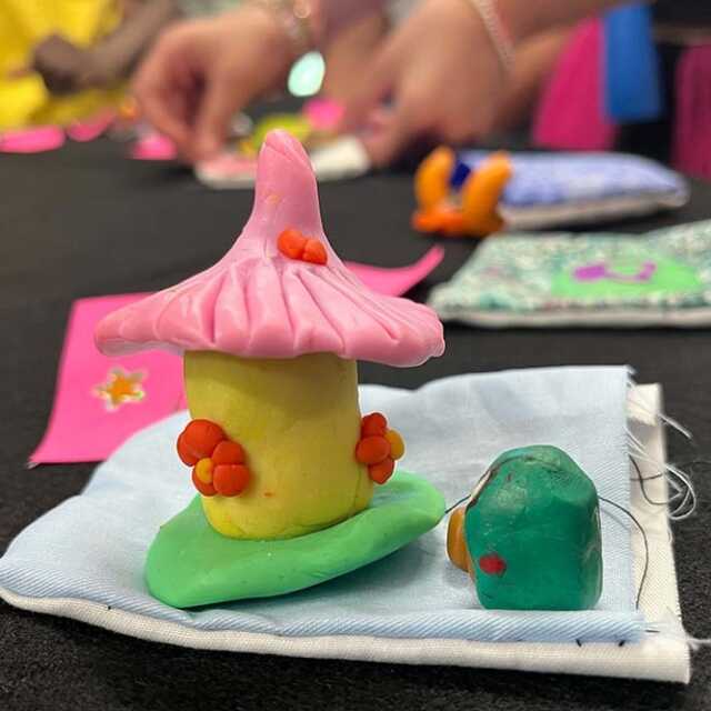 a small, yellow polymer clay fairy house with a pink, pointed roof sits next to a tiny green figure atop a hand-sewn cloth pocket. A students hands can be seen working in the background.