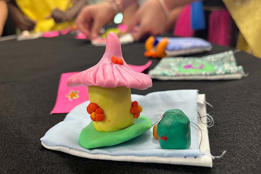 a small, yellow polymer clay fairy house with a pink, pointed roof sits next to a tiny green figure atop a hand-sewn cloth pocket. A students hands can be seen working in the background.