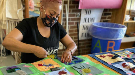 An adult wearing a floral fabric mask works on stitching a needle-felted fabric square to a larger piece of fabric