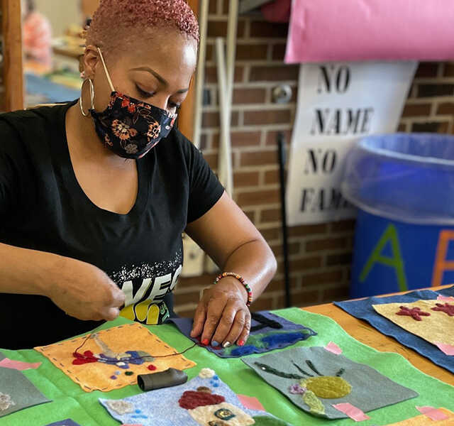An adult wearing a floral fabric mask works on stitching a needle-felted fabric square to a larger piece of fabric