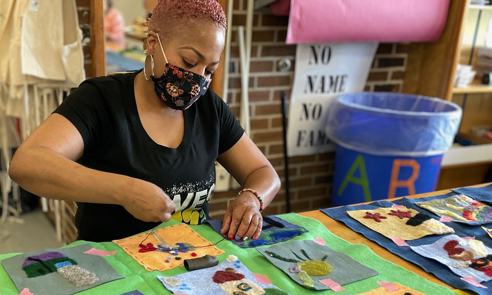 An adult wearing a floral fabric mask works on stitching a needle-felted fabric square to a larger piece of fabric