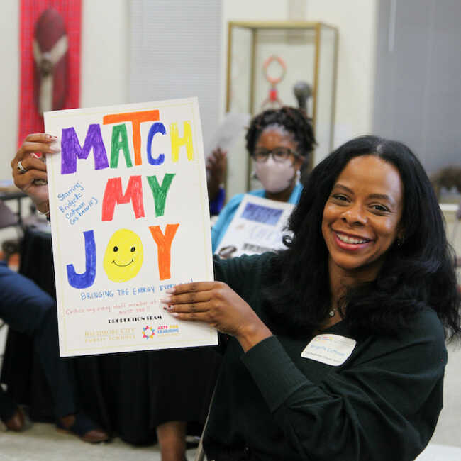 A woman dressed in black and wearing a name tag smiles as she holds a hand-drawn poster that says, "Match My Joy." A smiley face emoji stands in place of the letter O in the word 'joy.'