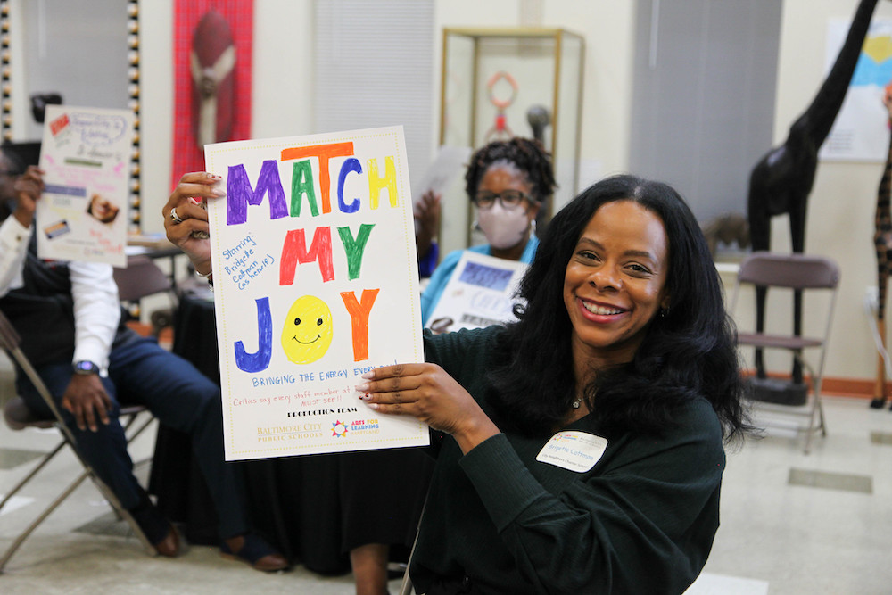 A woman dressed in black and wearing a name tag smiles as she holds a hand-drawn poster that says, "Match My Joy." A smiley face emoji stands in place of the letter O in the word 'joy.'