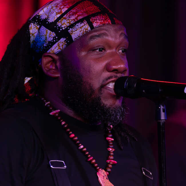 Mr. Word wears a patterned scarf wrapped around his hair and a chest-length, beaded necklace. He is speaking closely into a microphone onstage under red lights.