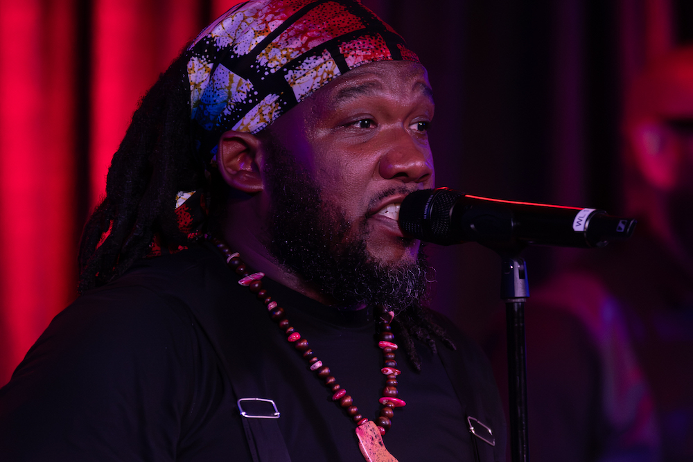 Mr. Word wears a patterned scarf wrapped around his hair and a chest-length, beaded necklace. He is speaking closely into a microphone onstage under red lights.