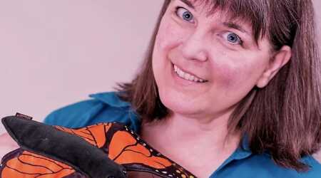 A smiling artist, her head tilted, with a large orange and black butterfly puppet, looks into the camera.