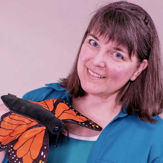 A smiling artist, her head tilted, with a large orange and black butterfly puppet, looks into the camera.
