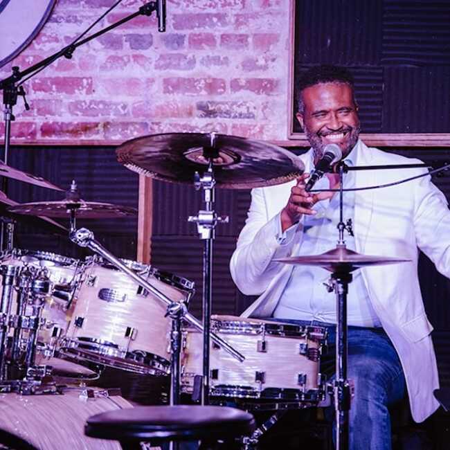 A drummer on stage in a white jacket and dress shirt is smiling, microphone in hand, sitting behind his drum set. A brick wall backs the stage.