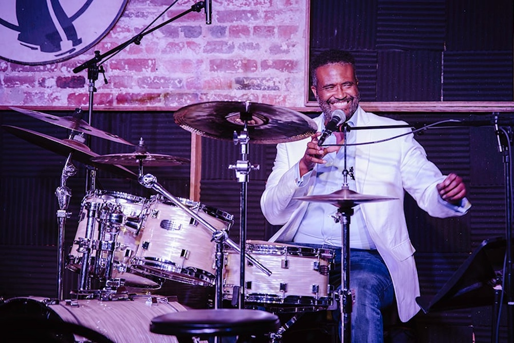 A drummer on stage in a white jacket and dress shirt is smiling, microphone in hand, sitting behind his drum set. A brick wall backs the stage.