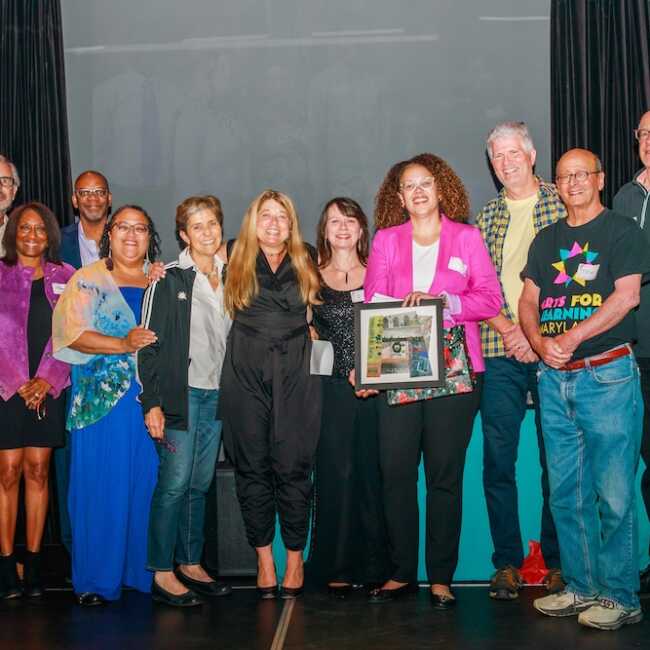 The Arts for Learning Maryland Board of Directors surround Stacie Sanders Evans on s stage. The group is smiling into the camera with Stacie in the middle. A board member in a bright pink blazer holds a framed photo collage stitched together by artist Katherine Dilworth.