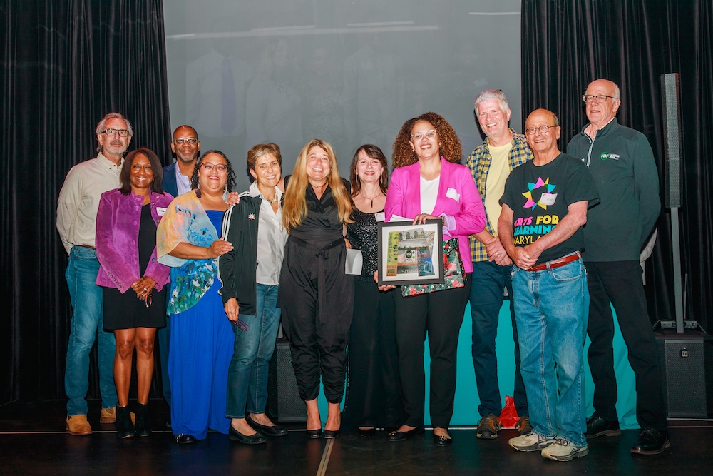 The Arts for Learning Maryland Board of Directors surround Stacie Sanders Evans on s stage. The group is smiling into the camera with Stacie in the middle. A board member in a bright pink blazer holds a framed photo collage stitched together by artist Katherine Dilworth.
