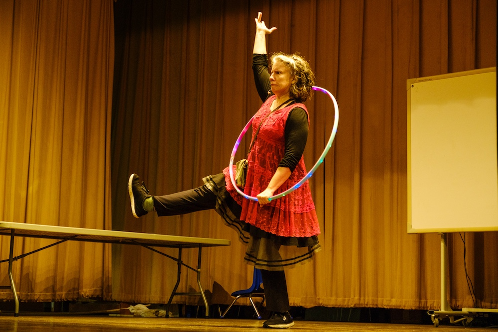 Michelle Minnick–wearing a red and black layered, loose fitting dress and stockings–stands on one leg as if frozen in an exaggerated march with one arm stretched high. She is on a school stage with a hula hoop criss-crossing her torso.