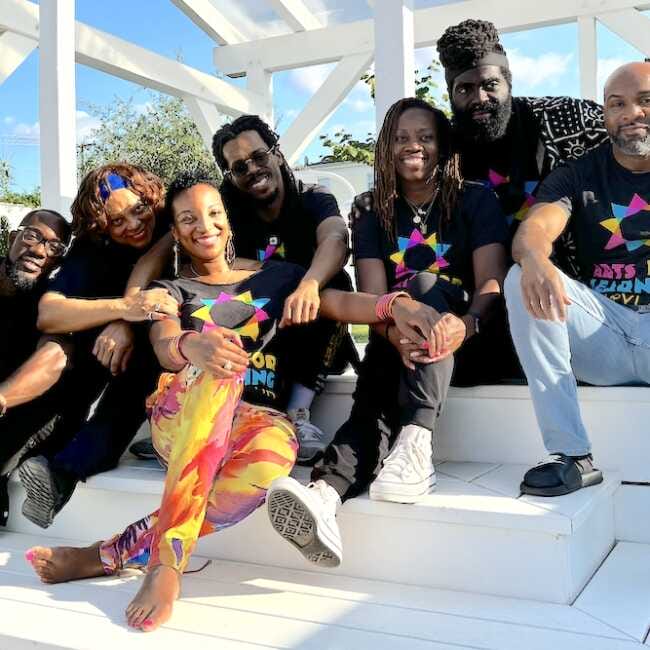 Blactastic performers Debra Mims, Baba Bomani, Jamaal “Mr. Root” Collier, Quynn Johnson, Drew Anderson, and Jessica “Culture Queen” Smith along with videographer Jimmy Powell gather in the shade of a white, wooden gazebo on a bright, sunny day. The group–in matching black t-shirts printed with Arts for Learning's bold cyan, magenta, and yellow logo–smiles into the camera.