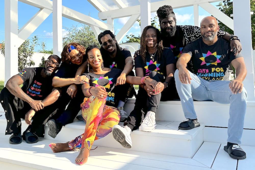 Blactastic performers Debra Mims, Baba Bomani, Jamaal “Mr. Root” Collier, Quynn Johnson, Drew Anderson, and Jessica “Culture Queen” Smith along with videographer Jimmy Powell gather in the shade of a white, wooden gazebo on a bright, sunny day. The group–in matching black t-shirts printed with Arts for Learning's bold cyan, magenta, and yellow logo–smiles into the camera.