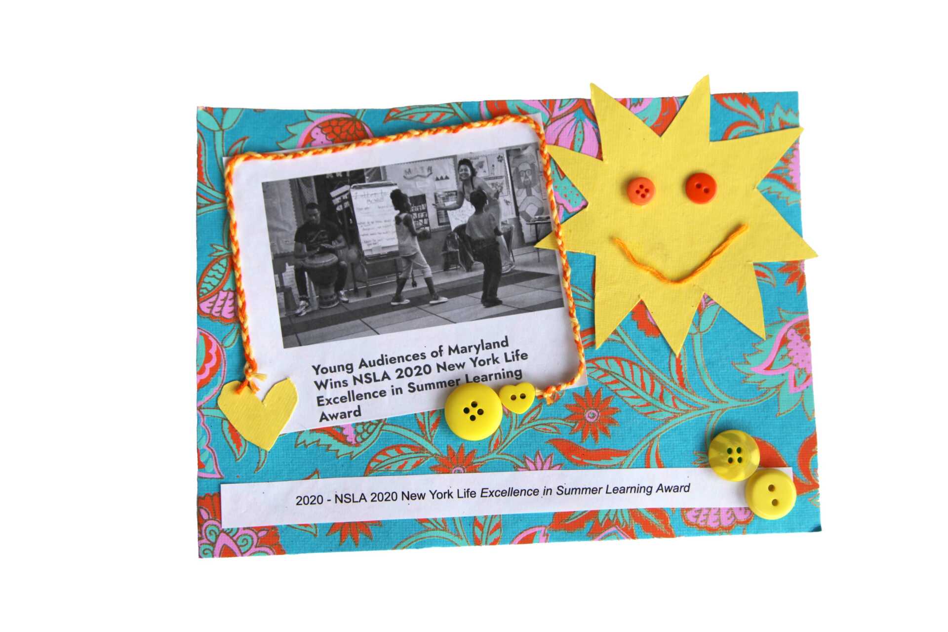 A smiling yellow sun-star beams in the corner over yellow buttons and hearts floating in a turquoise sky. An image of a teaching artist at the head of a classroom with the title,"Young Audiences of Maryland wins NSLA 2020 New York Life Excellence in Summer Learning Award."