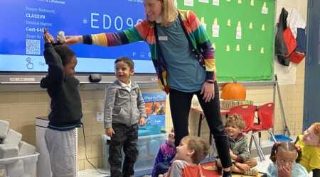 In a rainbow cardigan layered over a blue shirt and black leggings, Robin Neveu Brown stands an a small gathering of young school children. Her arm is outstretched with a hand positioning a tiny stuffed animal on top of a standing student's head.