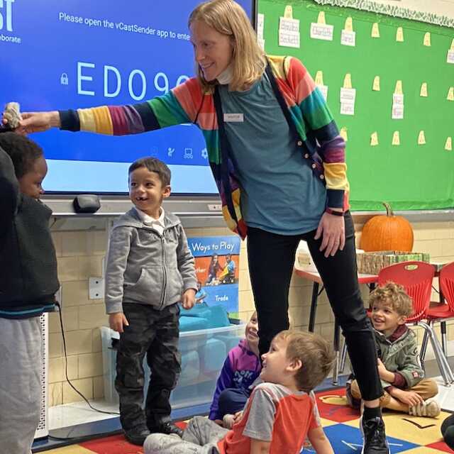 In a rainbow cardigan layered over a blue shirt and black leggings, Robin Neveu Brown stands an a small gathering of young school children. Her arm is outstretched with a hand positioning a tiny stuffed animal on top of a standing student's head.