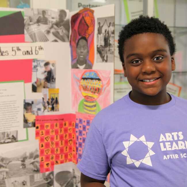 A child looking straight into the camera with a proud smile stands in front of a large trifold display collaged in children's art. They are wearing a lavender t-shirt that says, "Afer School Arts for Learning Academy."