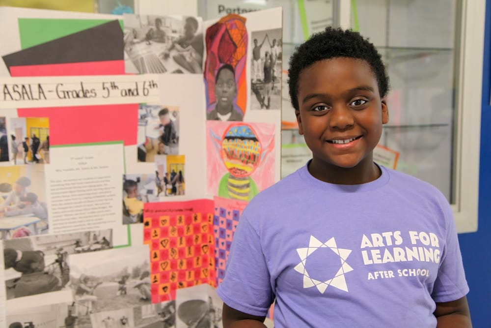 A child looking straight into the camera with a proud smile stands in front of a large trifold display collaged in children's art. They are wearing a lavender t-shirt that says, "Afer School Arts for Learning Academy."