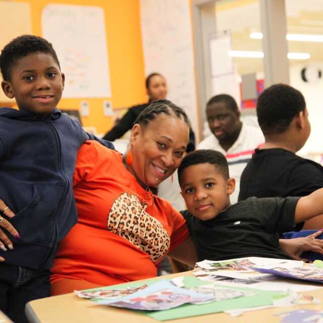 An adult in bright orange wearing a bright smile wraps their arms around two young students. They are at a table covered in paper materials in a bright gold and white classroom.