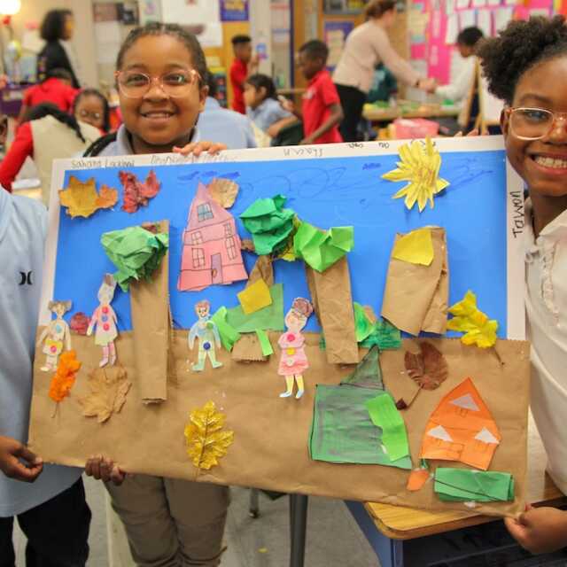 Smiling children hold up a collaborative construction paper collage in their classroom. Their artwork depicts a fall day in a neighborhood, complete with neighbors, leaves, houses, and even a pumpkin.