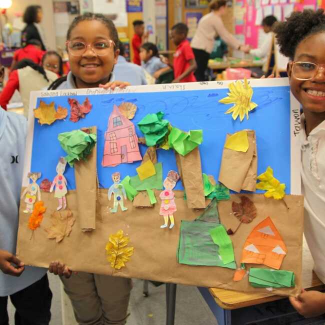 Smiling children hold up a collaborative construction paper collage in their classroom. Their artwork depicts a fall day in a neighborhood, complete with neighbors, leaves, houses, and even a pumpkin.