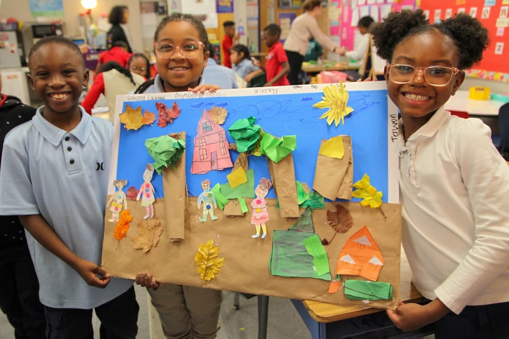 Smiling children hold up a collaborative construction paper collage in their classroom. Their artwork depicts a fall day in a neighborhood, complete with neighbors, leaves, houses, and even a pumpkin.