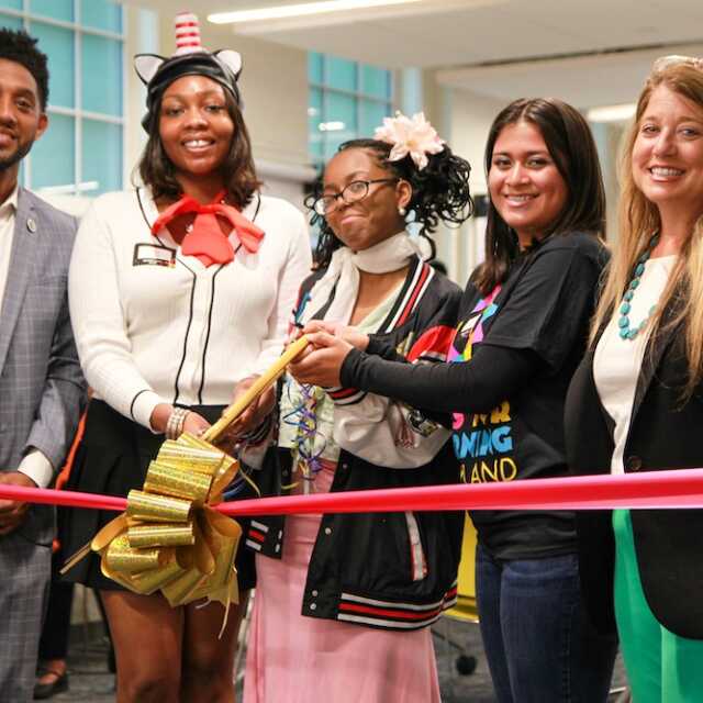 Baltimore Mayor Brandon Scott stands with Arts for Learning President and CEO Stacie Sanders Evans and three high-school students. All are smiling into the camera as the students brace a pair of giant golden scissors to cut a thick red ribbon and golden bow stretched out in front of them.