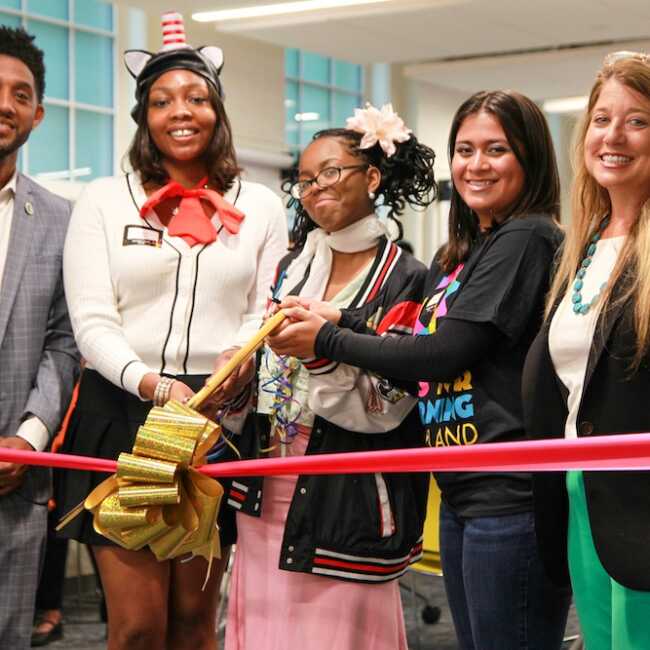 Baltimore Mayor Brandon Scott stands with Arts for Learning President and CEO Stacie Sanders Evans and three high-school students. All are smiling into the camera as the students brace a pair of giant golden scissors to cut a thick red ribbon and golden bow stretched out in front of them.