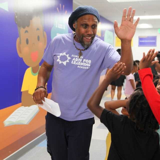 A teacher, smiling as he walks down the hallway, holds his hand up for high fives. A line of students are walking on the opposite side with hands up and ready.